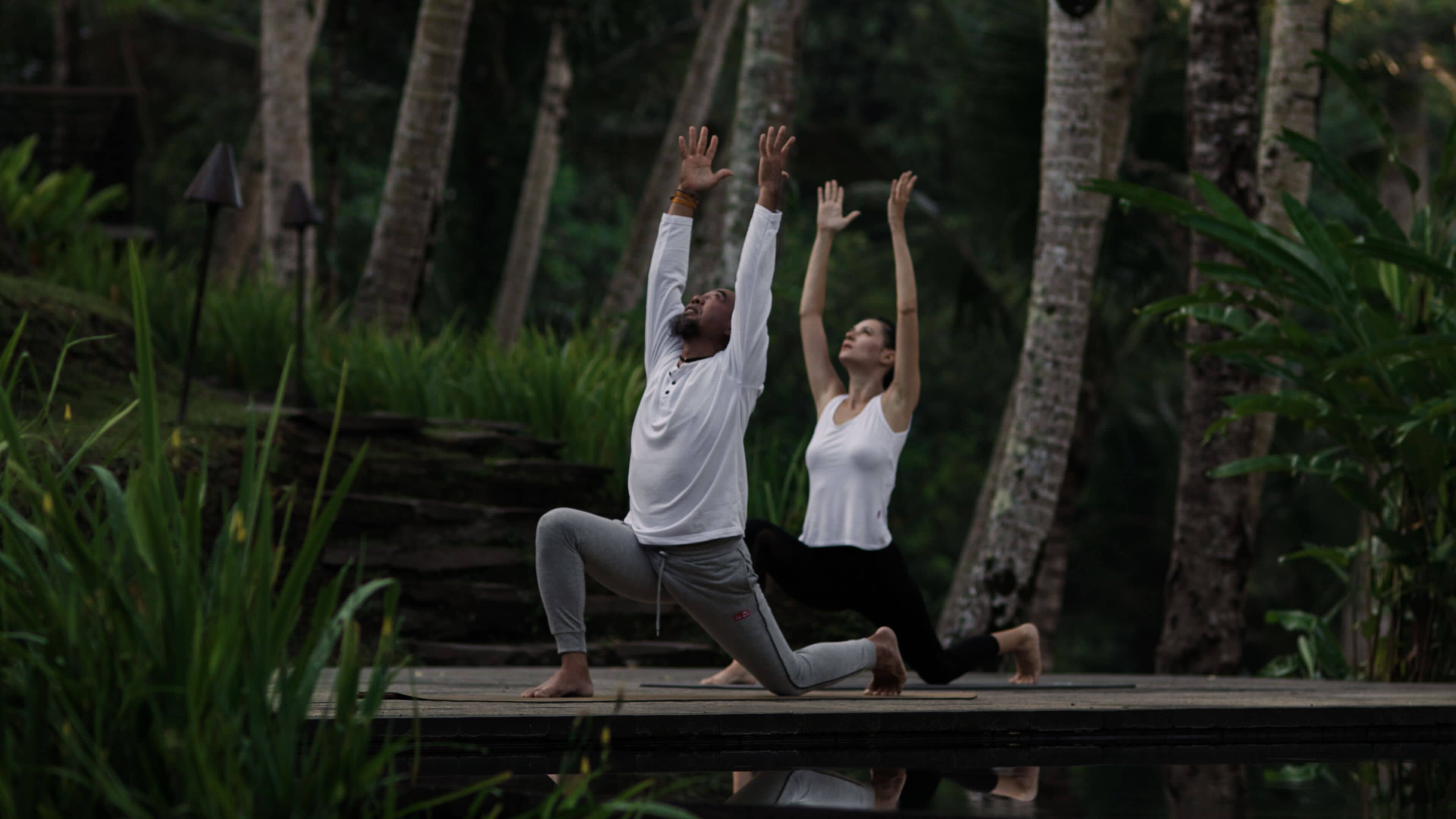 Deux personnes pratiquent le yoga sur une plateforme en bois, entourées de verdure.