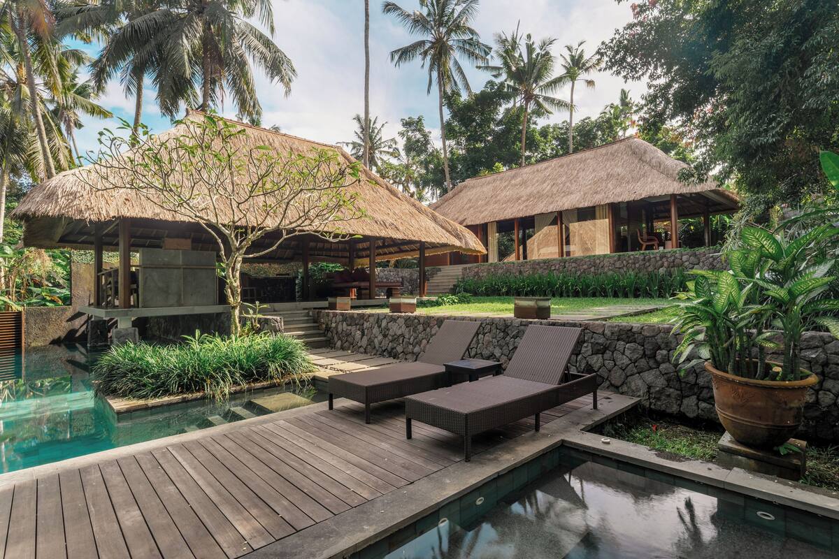 A poolside area with lounge chairs, a thatched-roof structure, and a stone wall.
