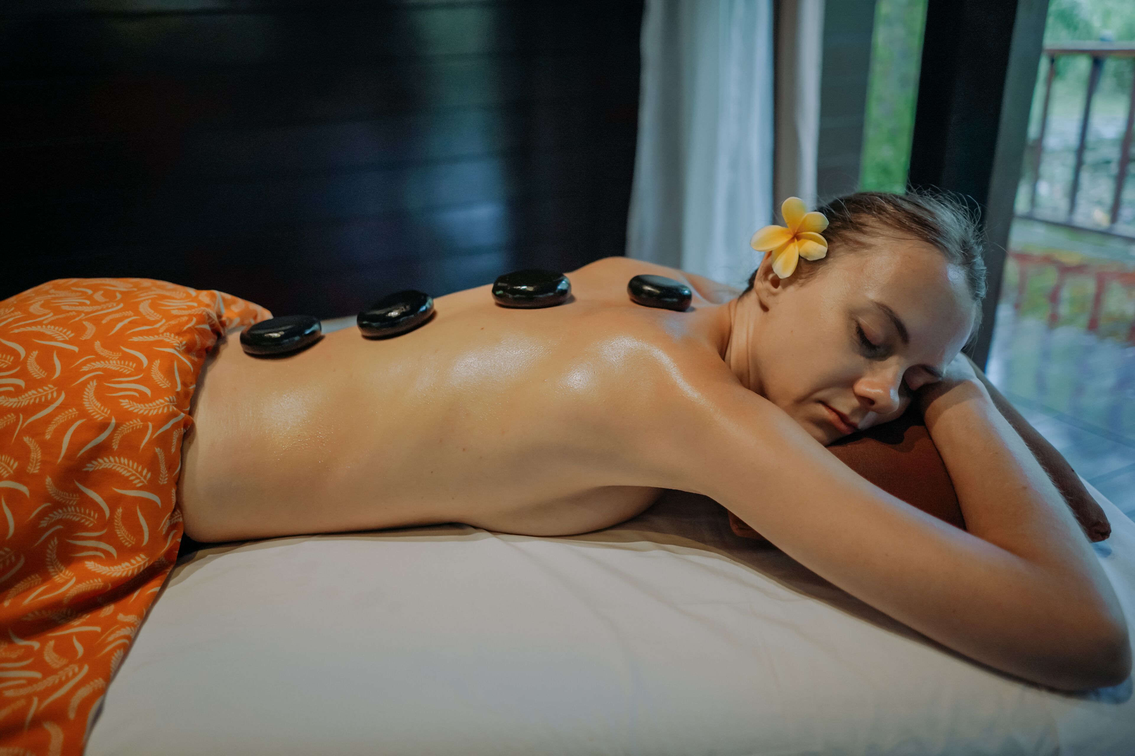 Una persona recibiendo un masaje con piedras calientes, con piedras colocadas sobre la espalda, acostada sobre una cama con una manta de estampado naranja.