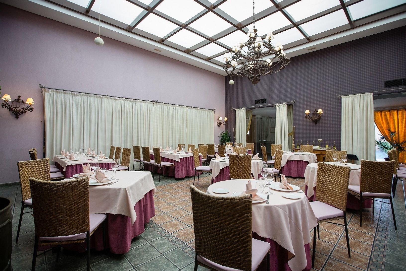 Un comedor con mesas dispuestas para el servicio, una araña y un amplio ventanal con cortinas.