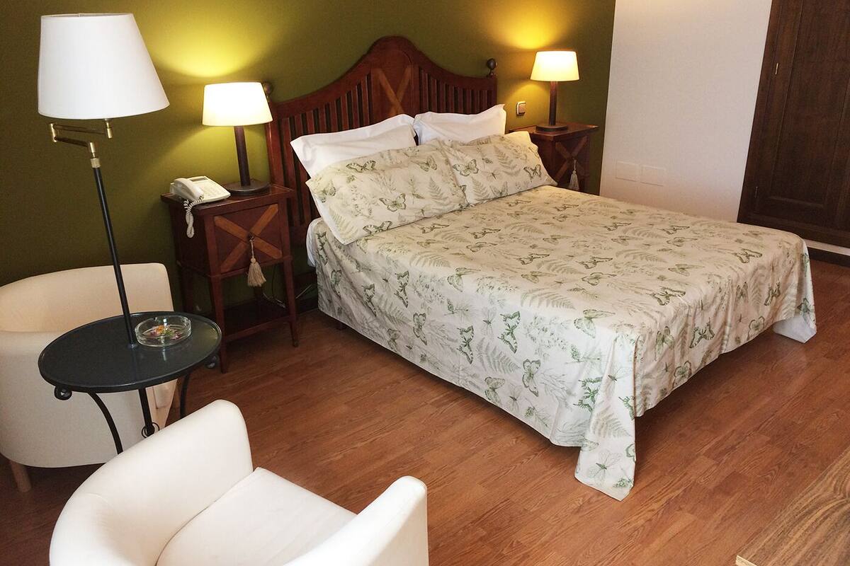 A neatly arranged bedroom with a bed, two bedside lamps, a telephone, a small table with a glass, and a white chair.