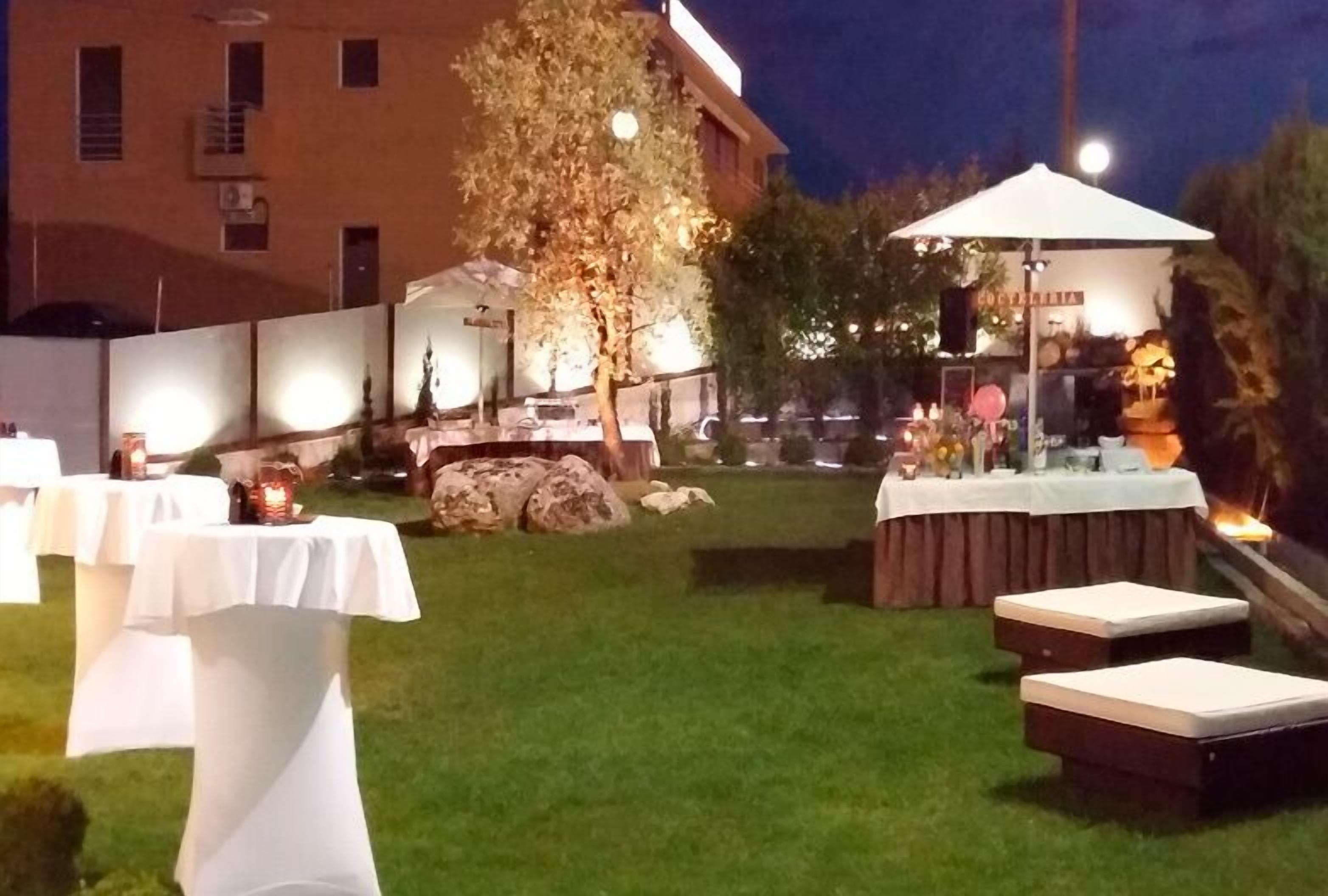 An outdoor patio area with tables and chairs, a small stage with a microphone, and a building in the background.