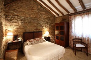 A bedroom with a stone wall, wooden beams, a bed with floral pillows, two bedside lamps, a wooden chair, and a door with glass panels. - El Añadio (Vilches)