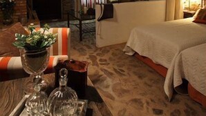 Une chambre confortable avec un mur en pierre, une grande fenêtre avec des rideaux, un lit recouvert d’une courtepointe blanche et une table de chevet sur laquelle se trouve un plateau de verres.
