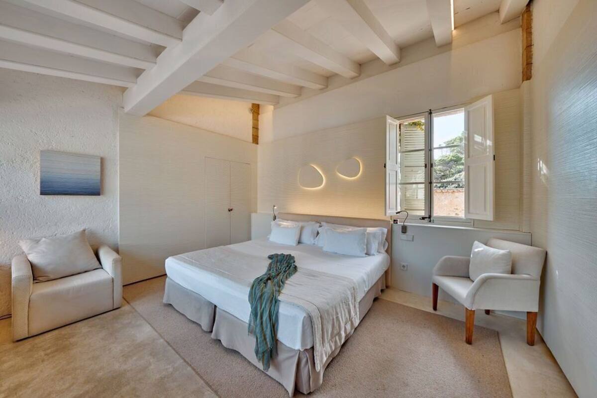 Un dormitorio con una cama grande, dos mesitas de noche, una ventana con persianas, un cuadro en la pared y un techo con vigas a la vista.