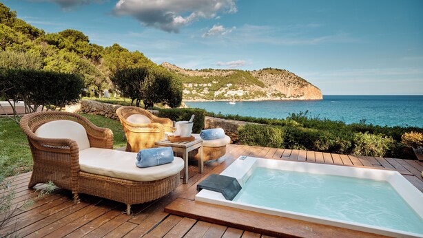 Una terraza de madera con mobiliario de mimbre, un jacuzzi y vistas al mar y colinas.