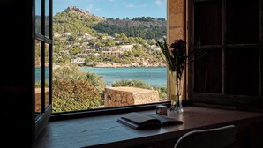 Una habitación con un ventanal que ofrece vistas a un pueblo costero, un banco de piedra, un florero con flores y un libro en el alféizar.