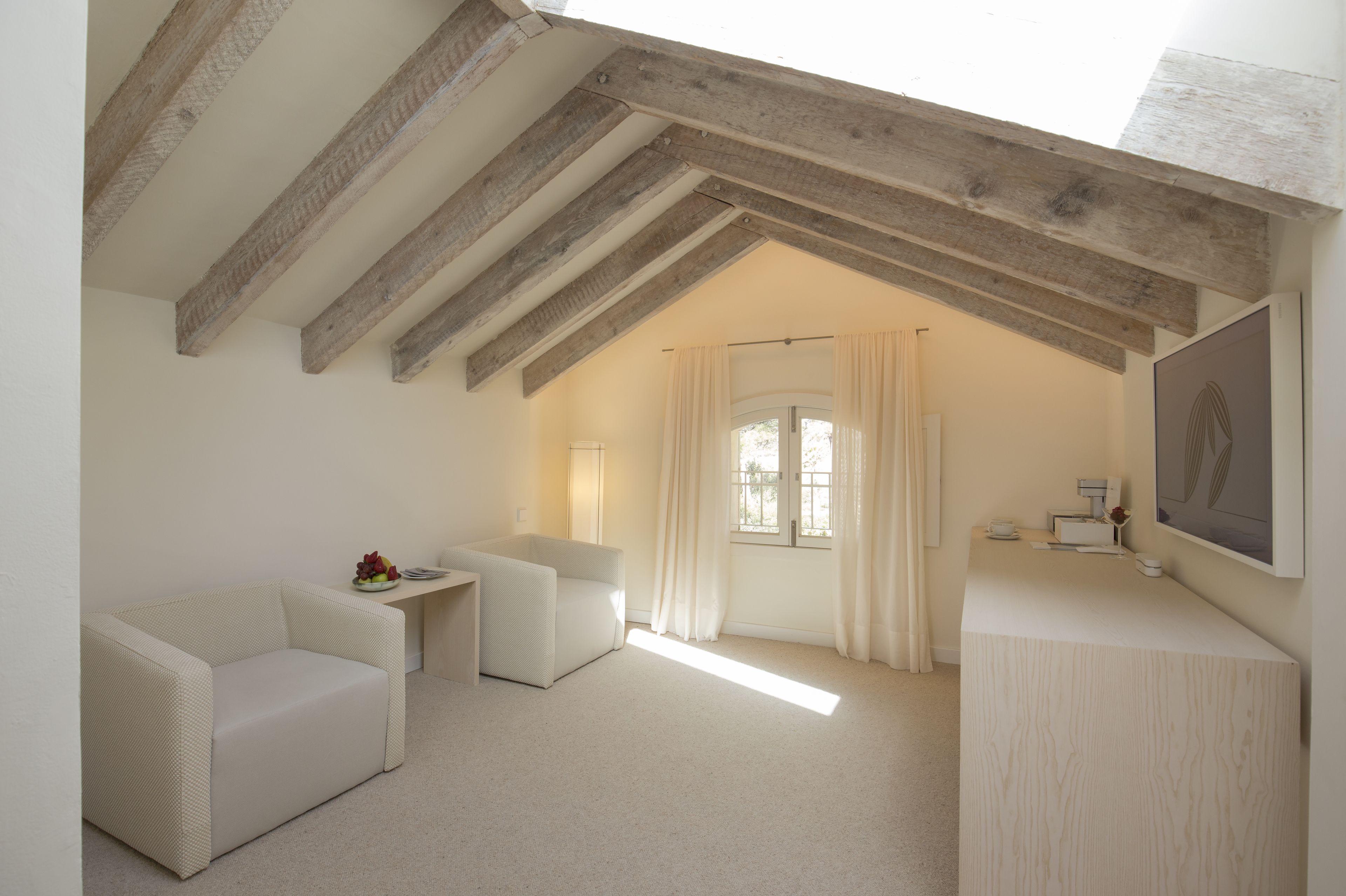 A room with a sloped ceiling, a sofa, a small table, a desk, and a window with curtains.
