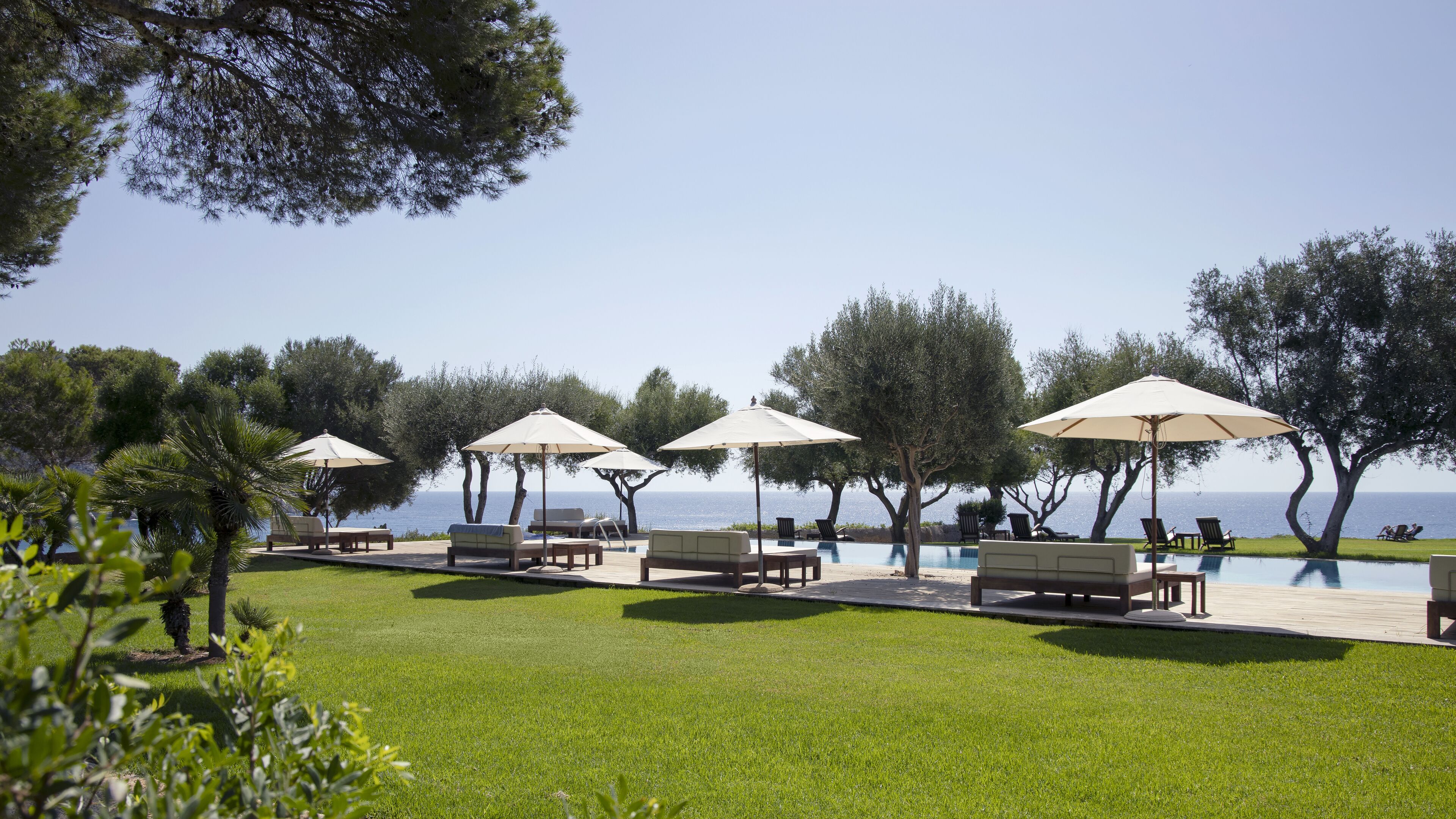 A poolside area with lounge chairs and umbrellas, surrounded by trees and a view of the sea.