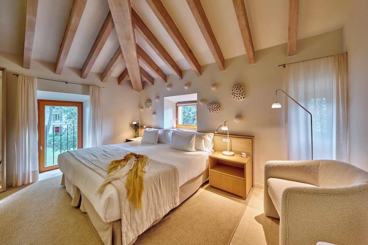 Un dormitorio con una cama grande, vigas de madera, una mesita de noche, una lámpara y vistas al exterior.