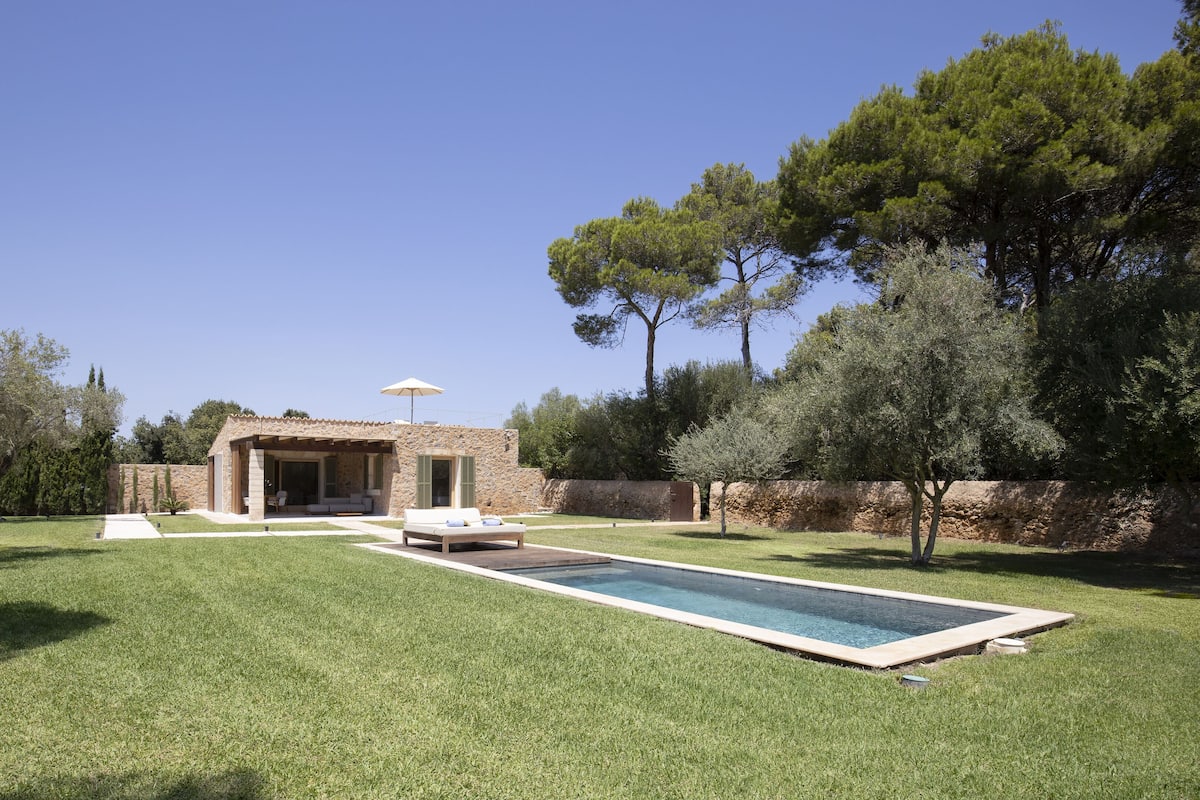 Una villa moderna con piscina, mobiliario exterior y un muro de piedra.