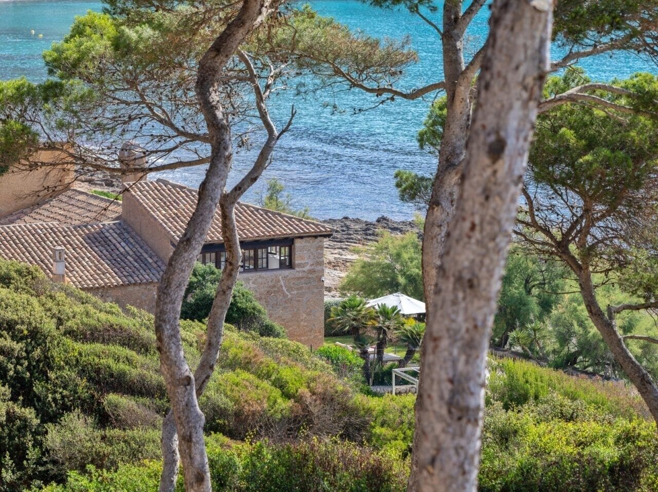 A stone house with a tiled roof overlooking a coastal area with trees and a view of the sea.