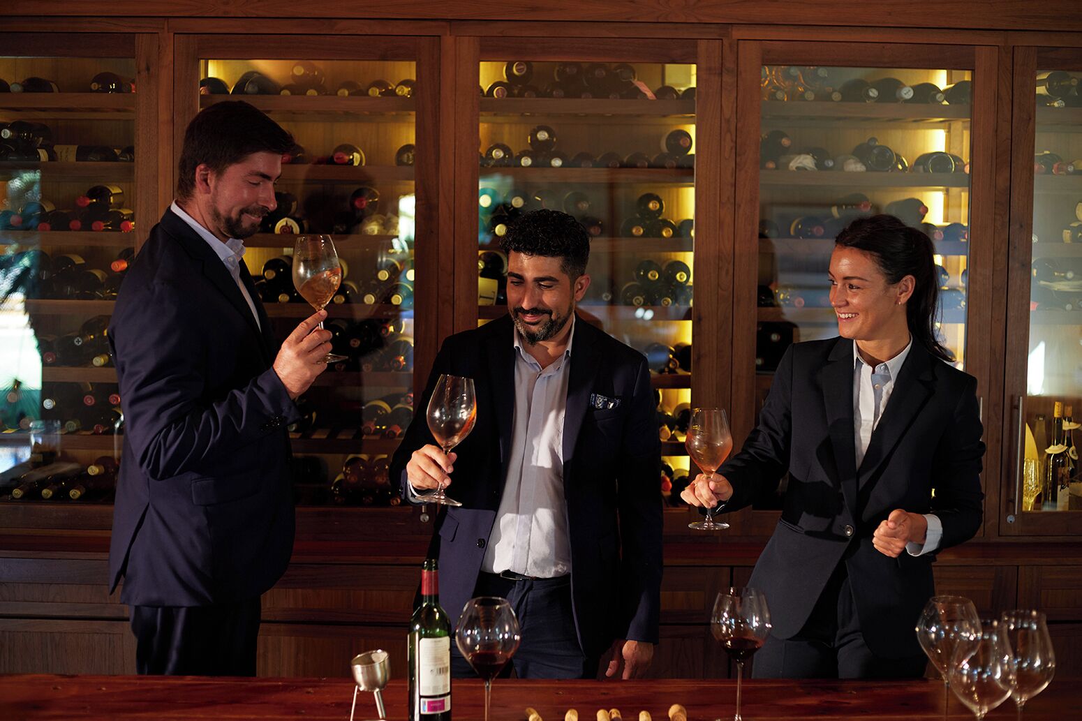Three people toasting with wine glasses at a bar with a wine bottle and glasses on the counter.