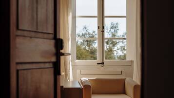 Una habitación con puerta de madera, ventana con vista a árboles y un sillón color crema.