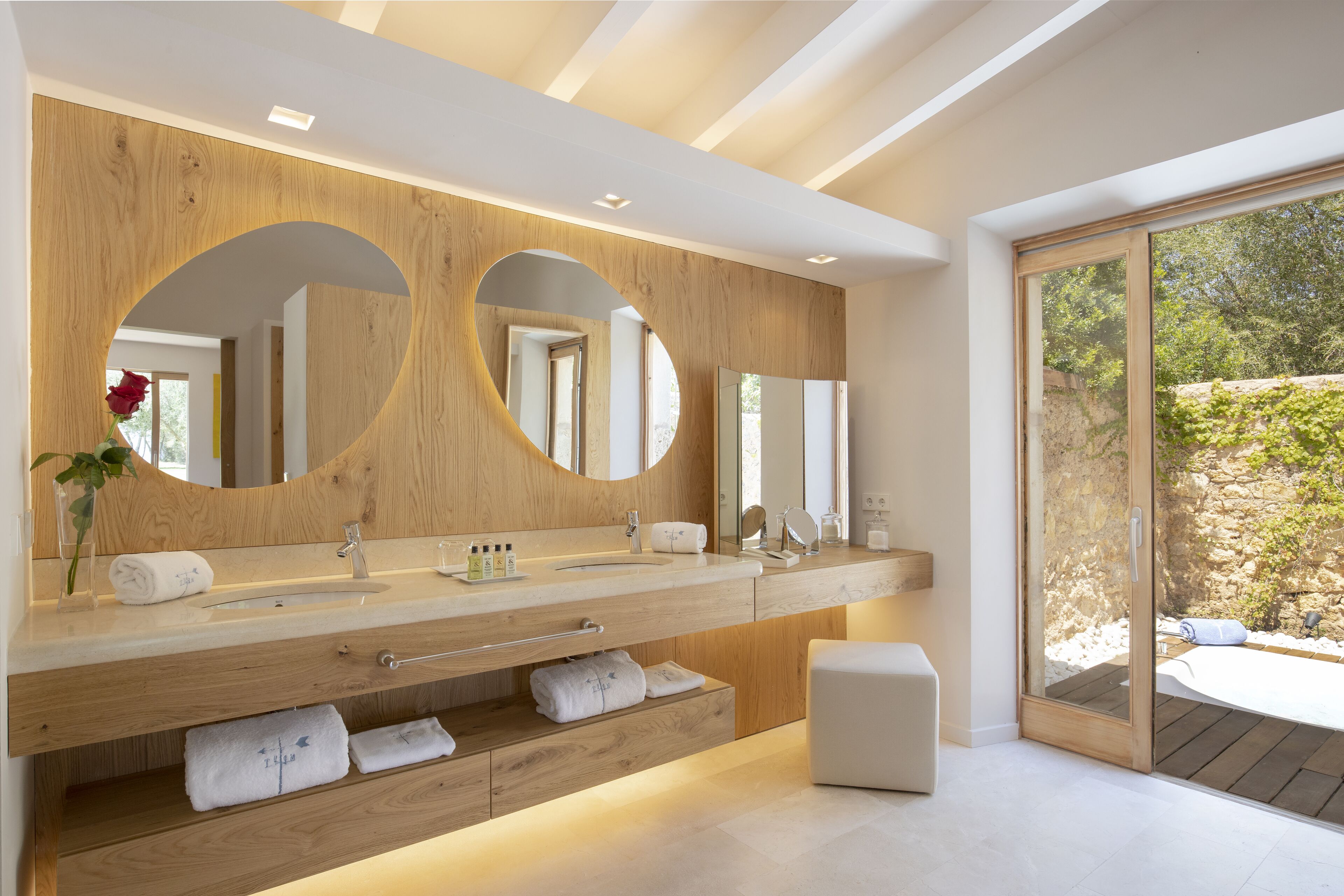 A modern bathroom with a wooden vanity, dual mirrors, and a large window offering a view of a garden.