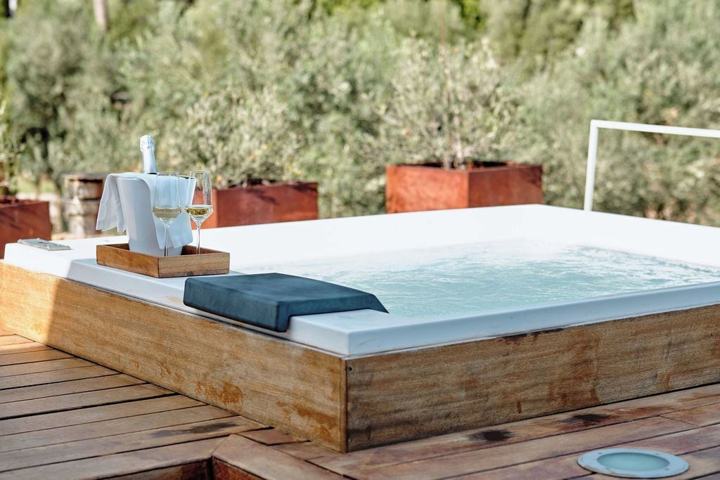 Een houten jacuzzi met een witte rand, een handdoek en een glas witte wijn op een houten terras.