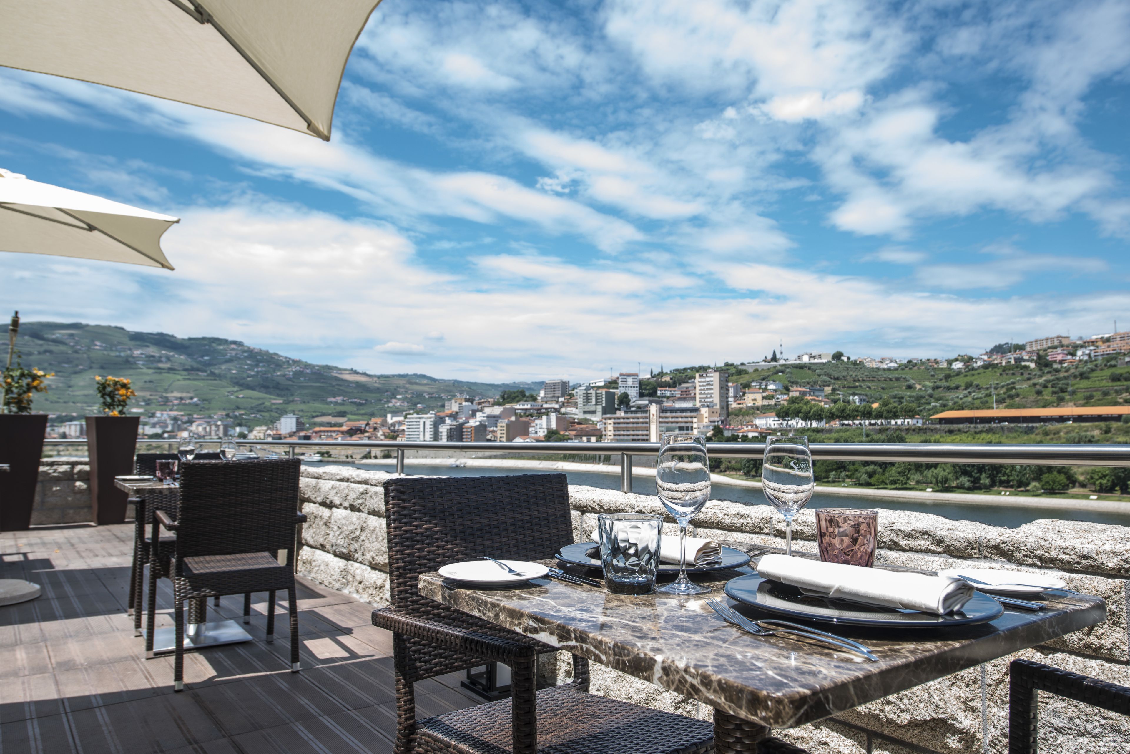 An outdoor dining area with wicker chairs and a table set for two, overlooking a cityscape with hills in the background.