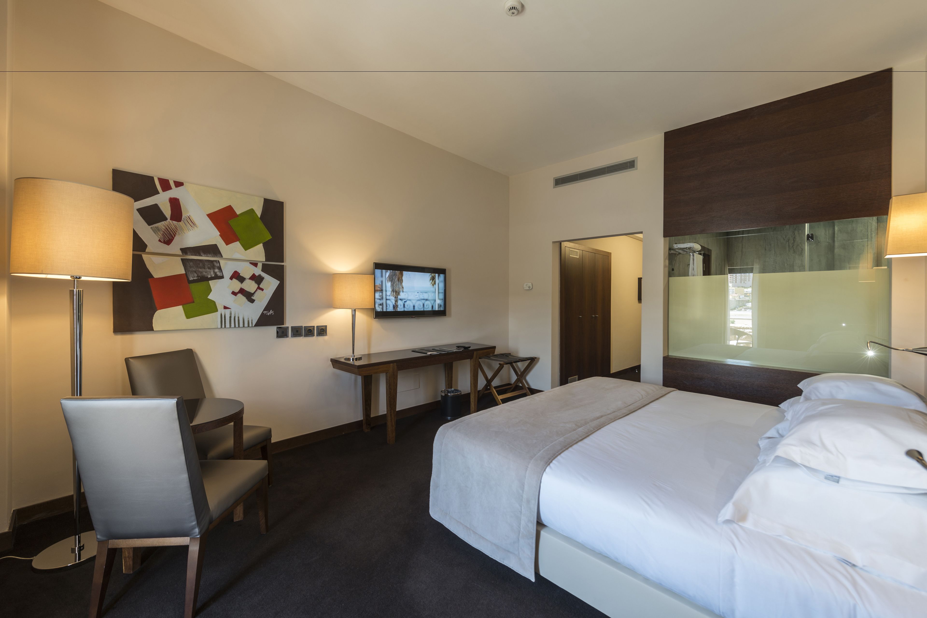 A hotel room with a bed, desk, chair, TV, and artwork on the wall.