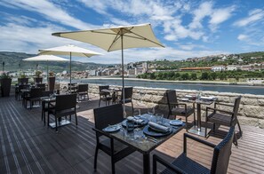 An outdoor dining area with tables and chairs, large umbrellas, and a scenic view of a river and town in the background. - Vila Gale Collection Douro Hotel (Lamego)