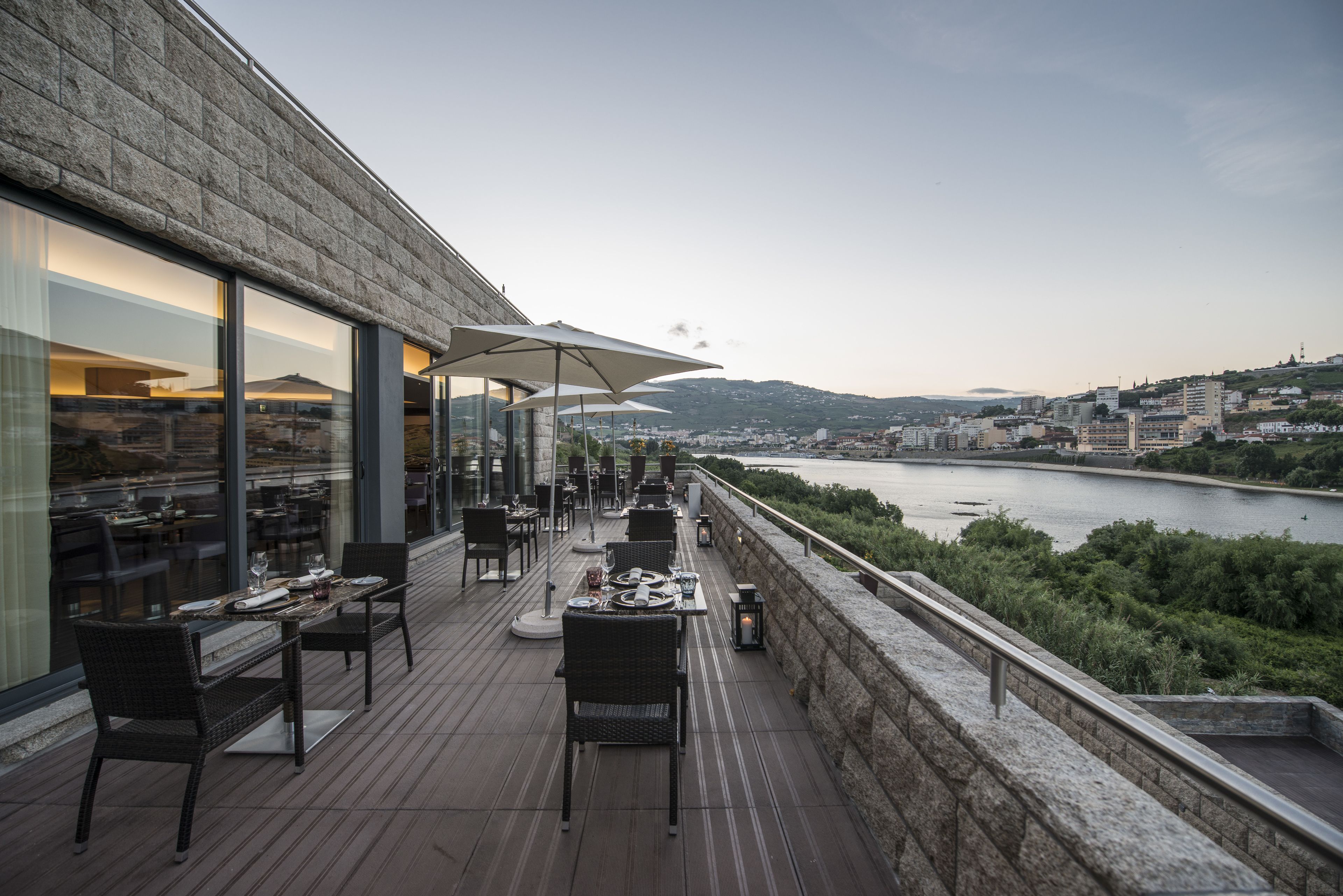 Uma área de refeições ao ar livre com mesas e cadeiras, com vista para um rio e o horizonte da cidade.