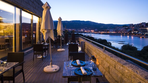 An outdoor dining area with tables and chairs, overlooking a river and cityscape. - Vila Gale Collection Douro Hotel (Lamego)