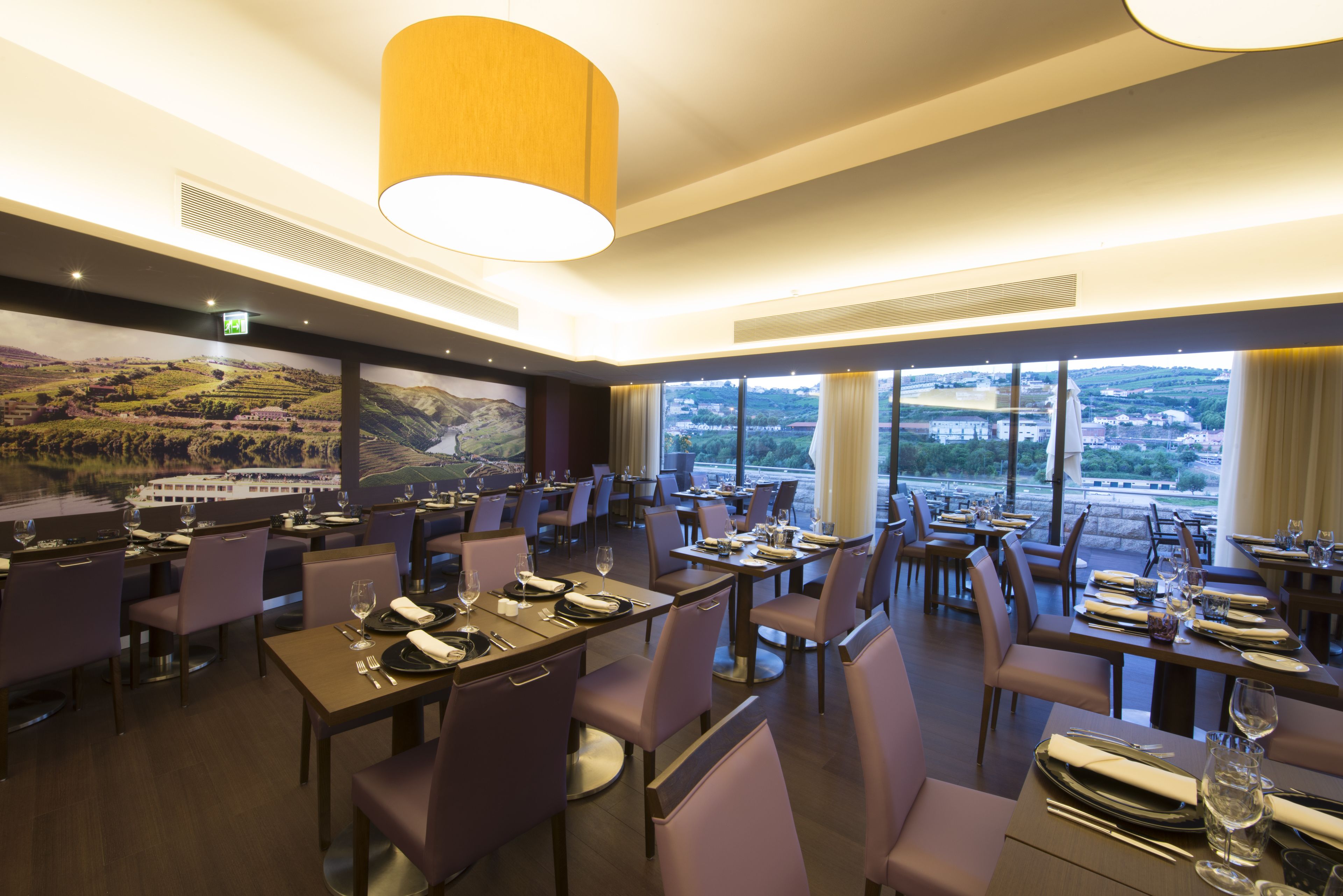 Un restaurante con varias mesas y sillas, amplias ventanas con vista a un paisaje, y una lámpara colgante grande.