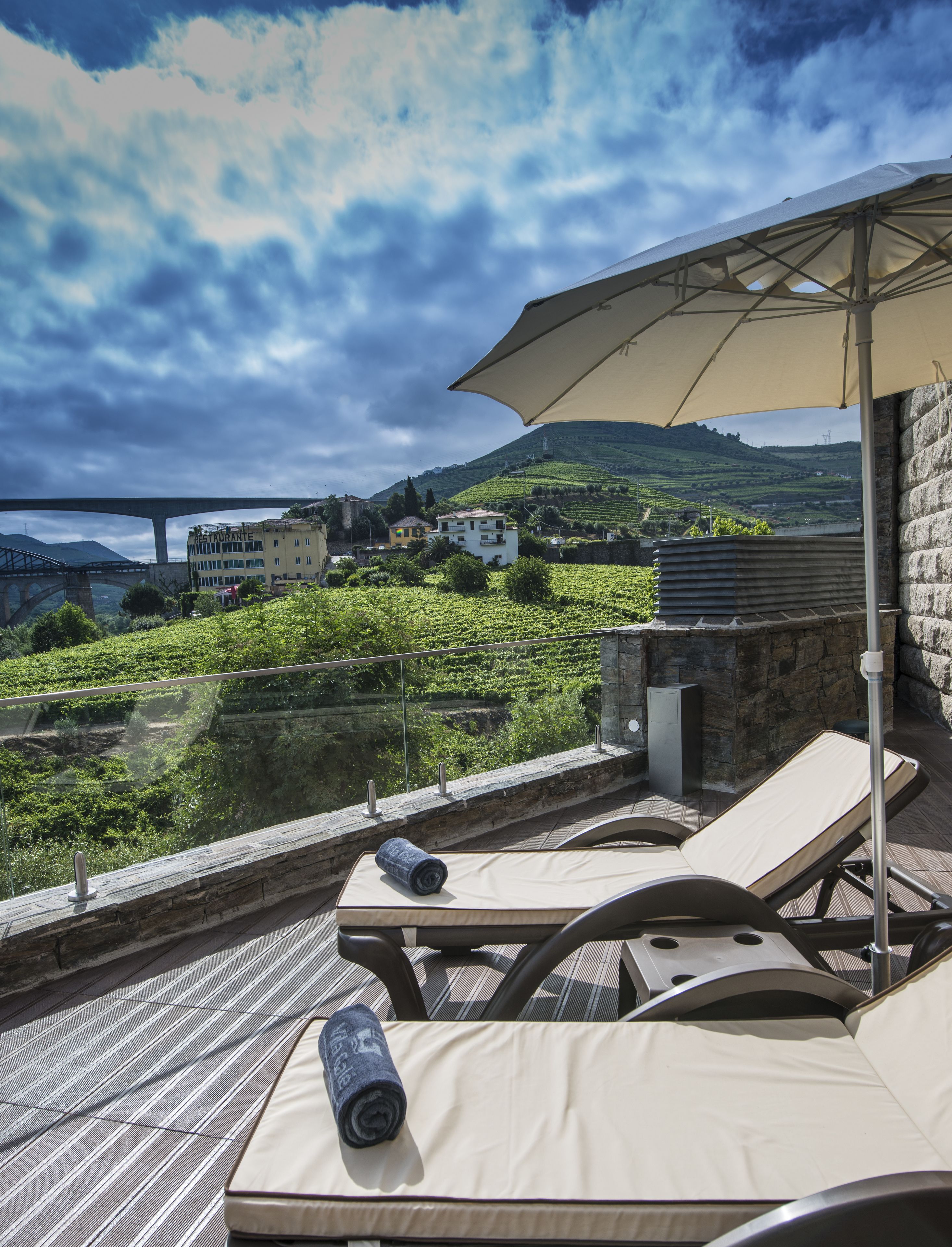 Um terraço com espreguiçadeiras, uma mesa e um grande guarda-sol. É visível uma vista para um vinhedo e uma ponte ao fundo.