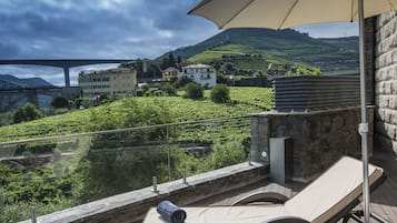 Um terraço com espreguiçadeiras, uma mesa e um grande guarda-sol. É visível uma vista para um vinhedo e uma ponte ao fundo.