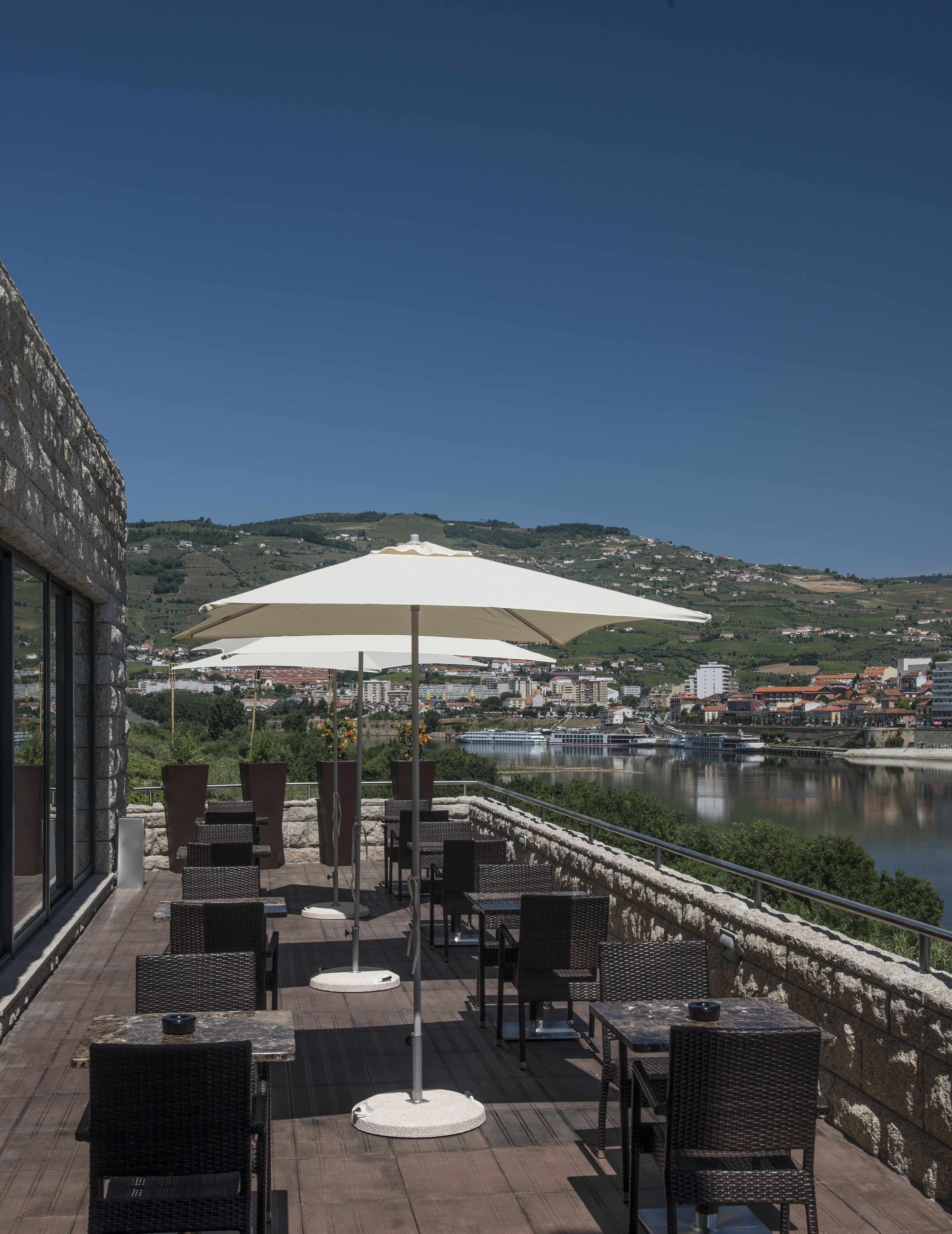 An outdoor terrace with wicker furniture, white umbrellas, and a view of a river and town in the background.