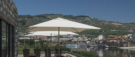 An outdoor terrace with wicker furniture, white umbrellas, and a view of a river and town in the background.