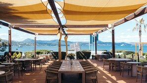 Een buitenterras met houten vloer, tafels en stoelen, met uitzicht op een waterpartij en bergen.