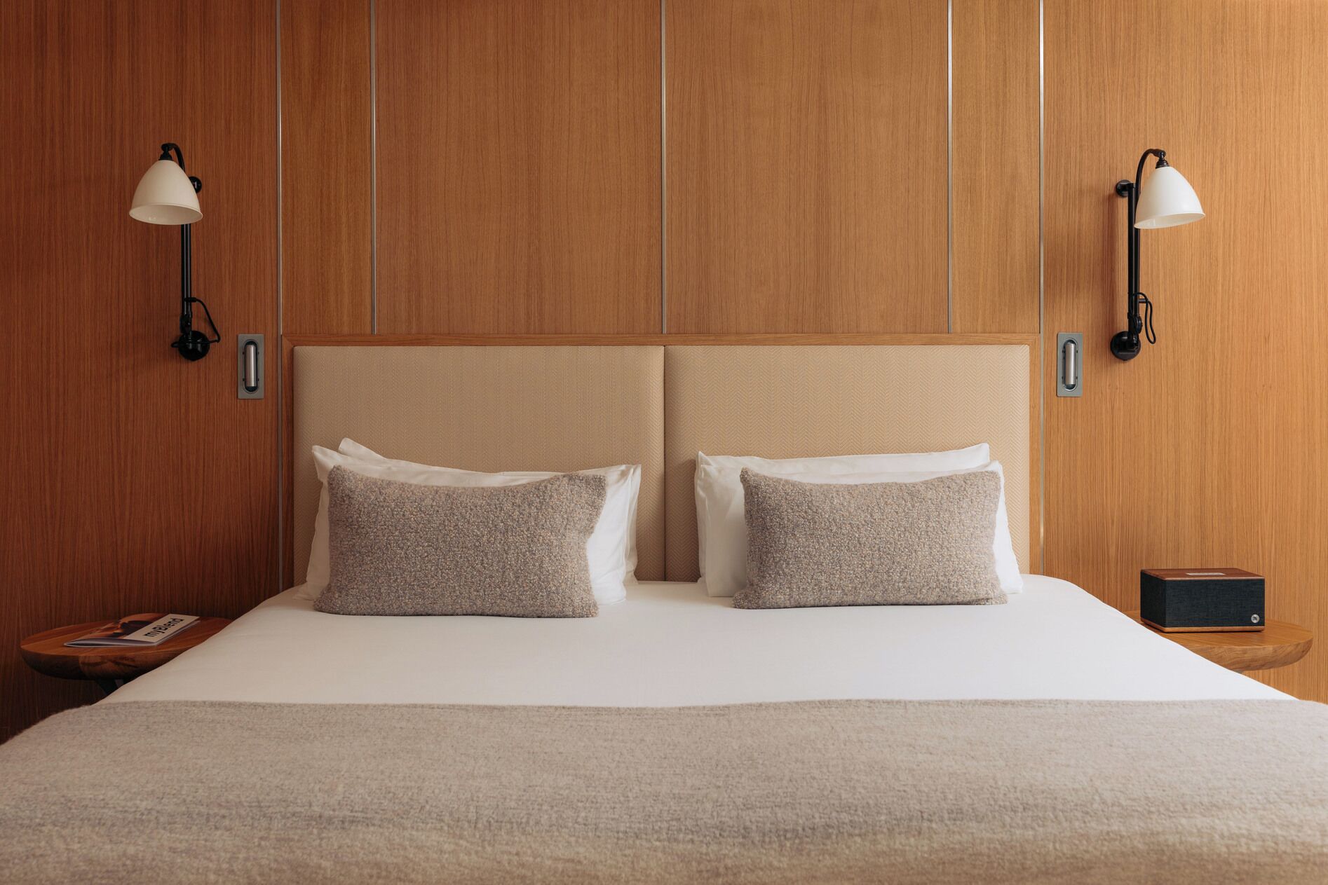 Une chambre d’hôtel avec un grand lit, deux lampes fixées au mur et une table de chevet avec un livre.