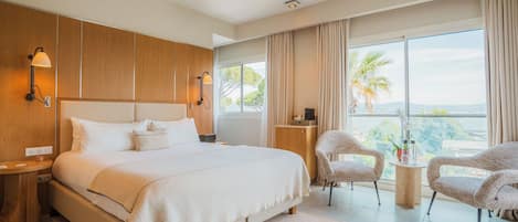 A modern hotel room with a large bed, wooden panel walls, a small desk, two chairs, and a view of the outdoors.