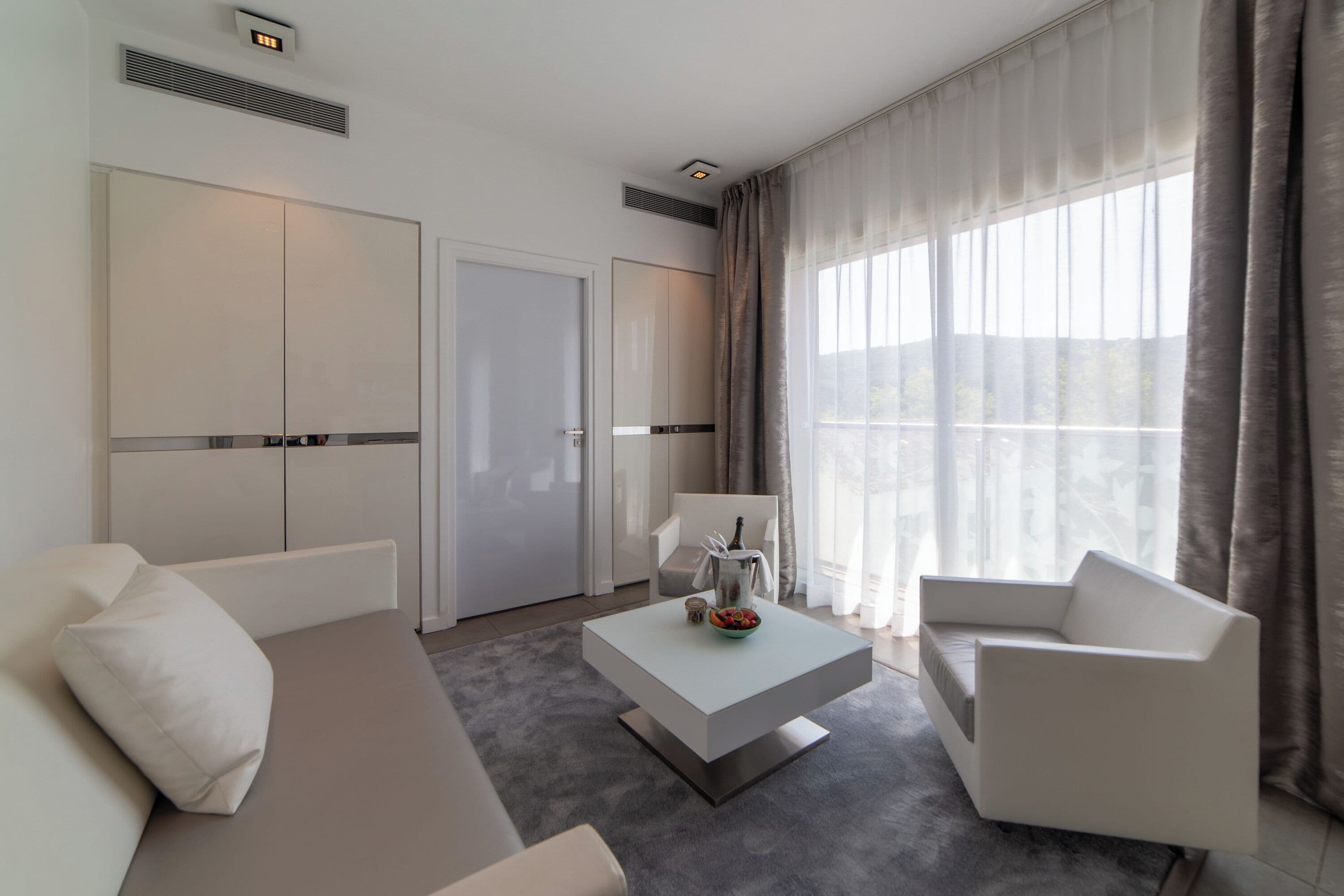 Ett modernt hotellrum med en soffa, ett soffbord, en stol och ett stort fönster med gardiner.