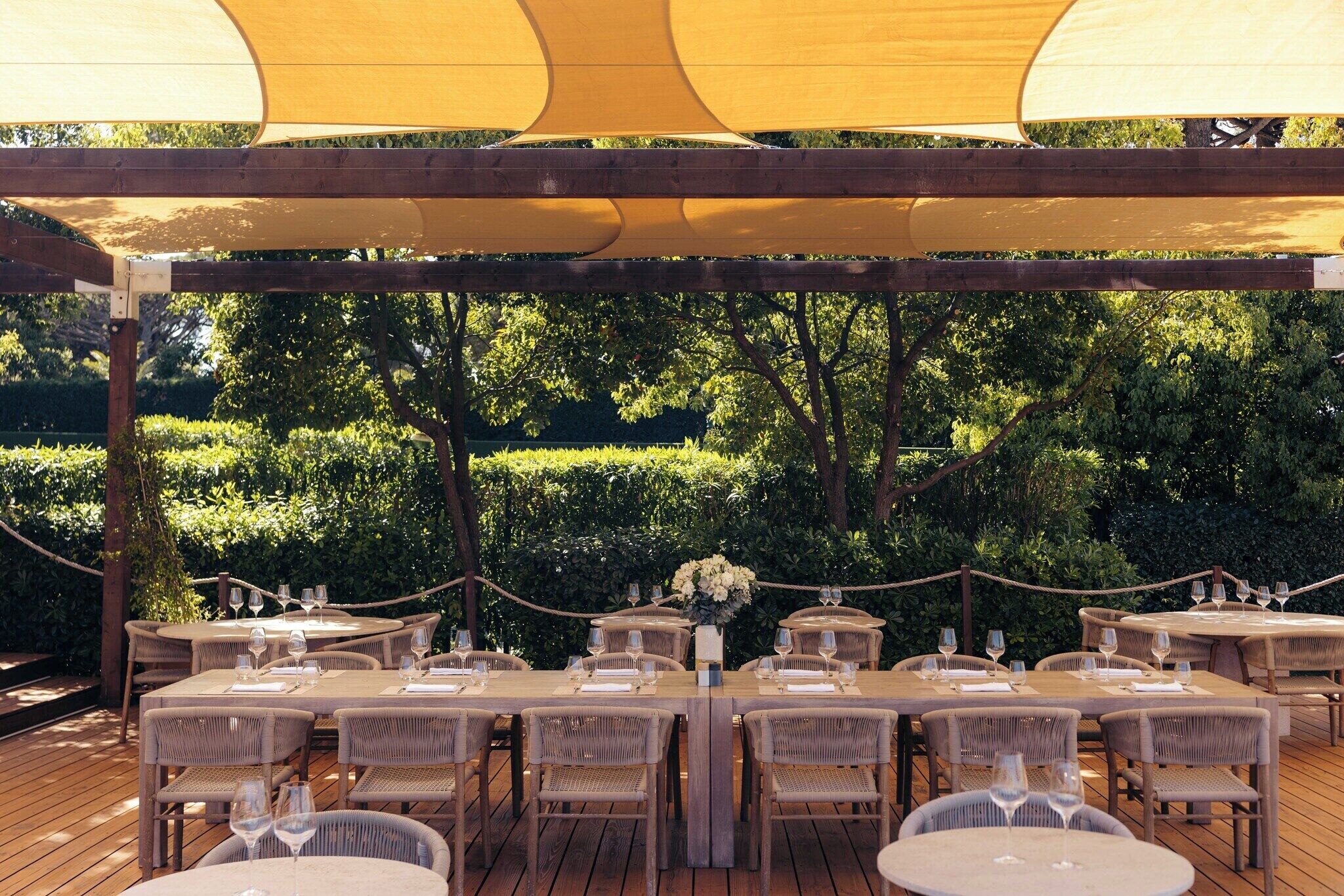 Un área de comedor al aire libre con mesas redondas y sillas de mimbre, rodeada por una terraza de madera y un dosel amarillo.