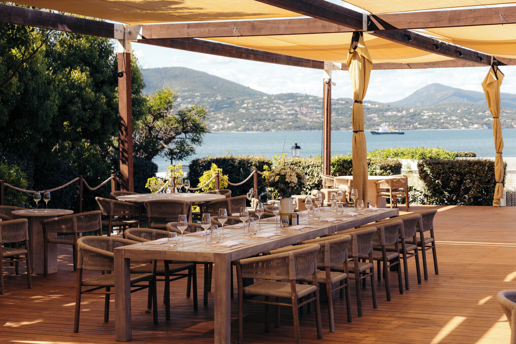 An outdoor dining area with wooden tables and chairs, a view of the sea and mountains, and a rope barrier.