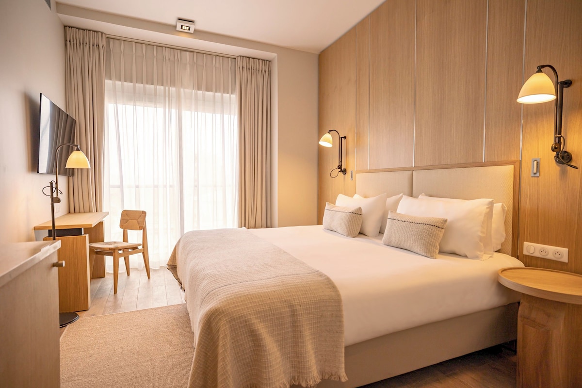 A hotel room with a bed, a desk, a chair, a TV, and two wall-mounted lamps.