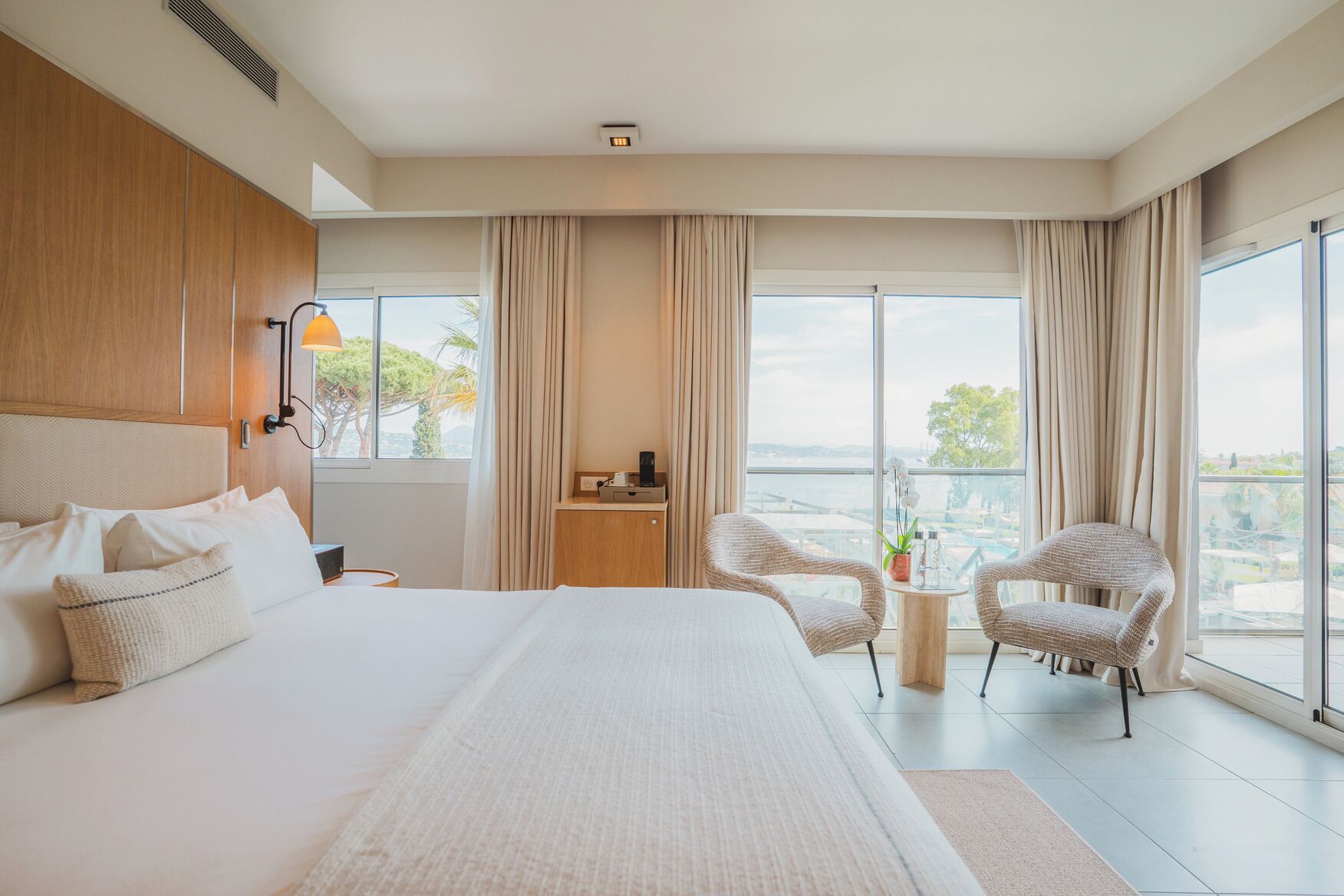 Een moderne hotelkamer met een groot bed, twee rieten stoelen, een klein tafeltje en uitzicht op de stad door grote ramen.
