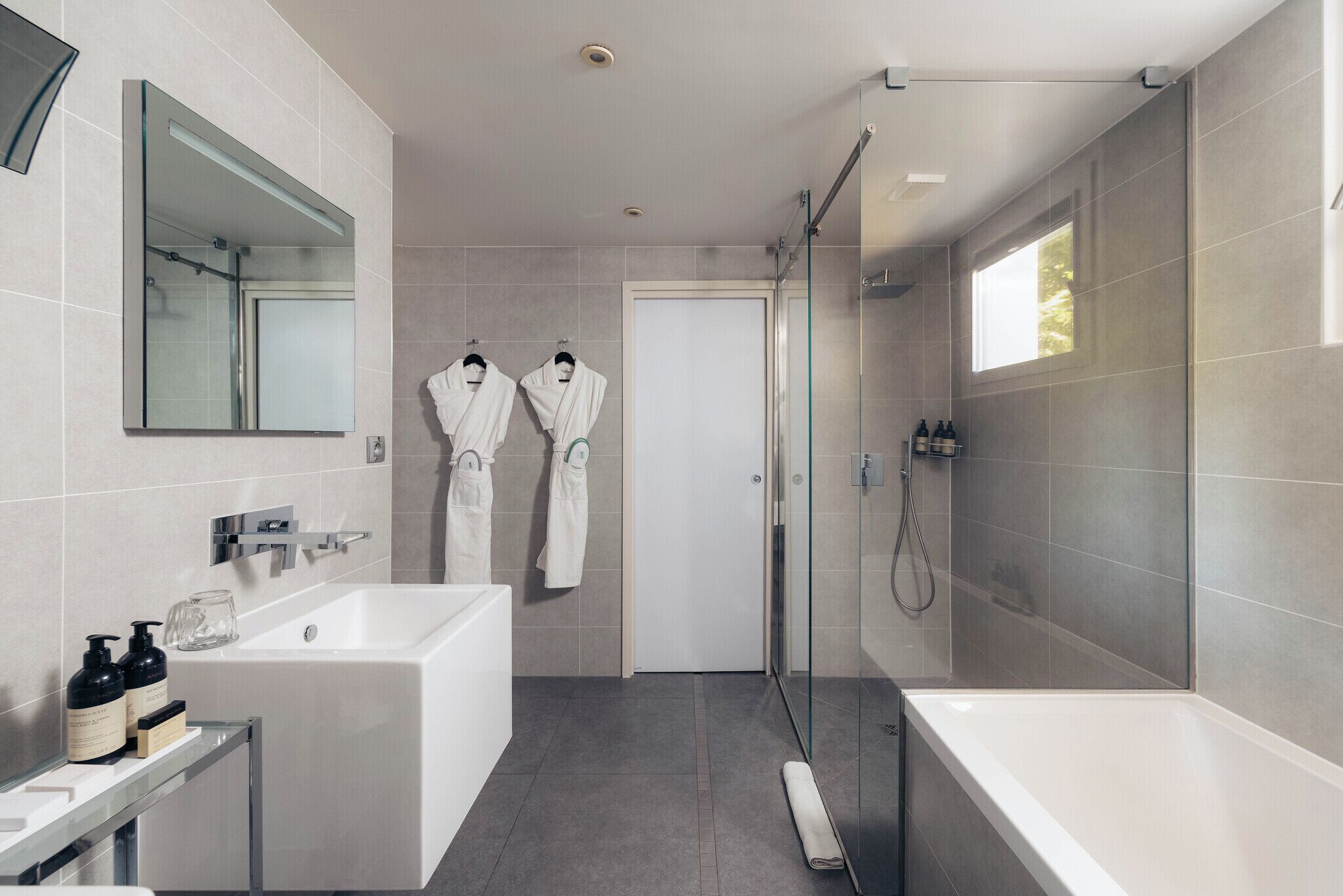 Un baño moderno con bañera, ducha con mampara de vidrio y dos albornoces blancos colgados en ganchos.