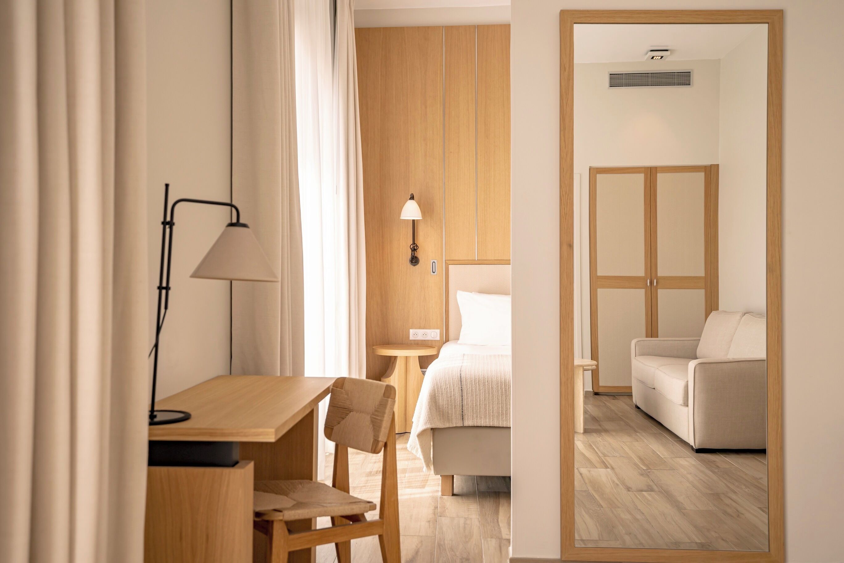 A hotel room with a wooden desk, chair, bedside table, lamp, and a bed with a white headboard.