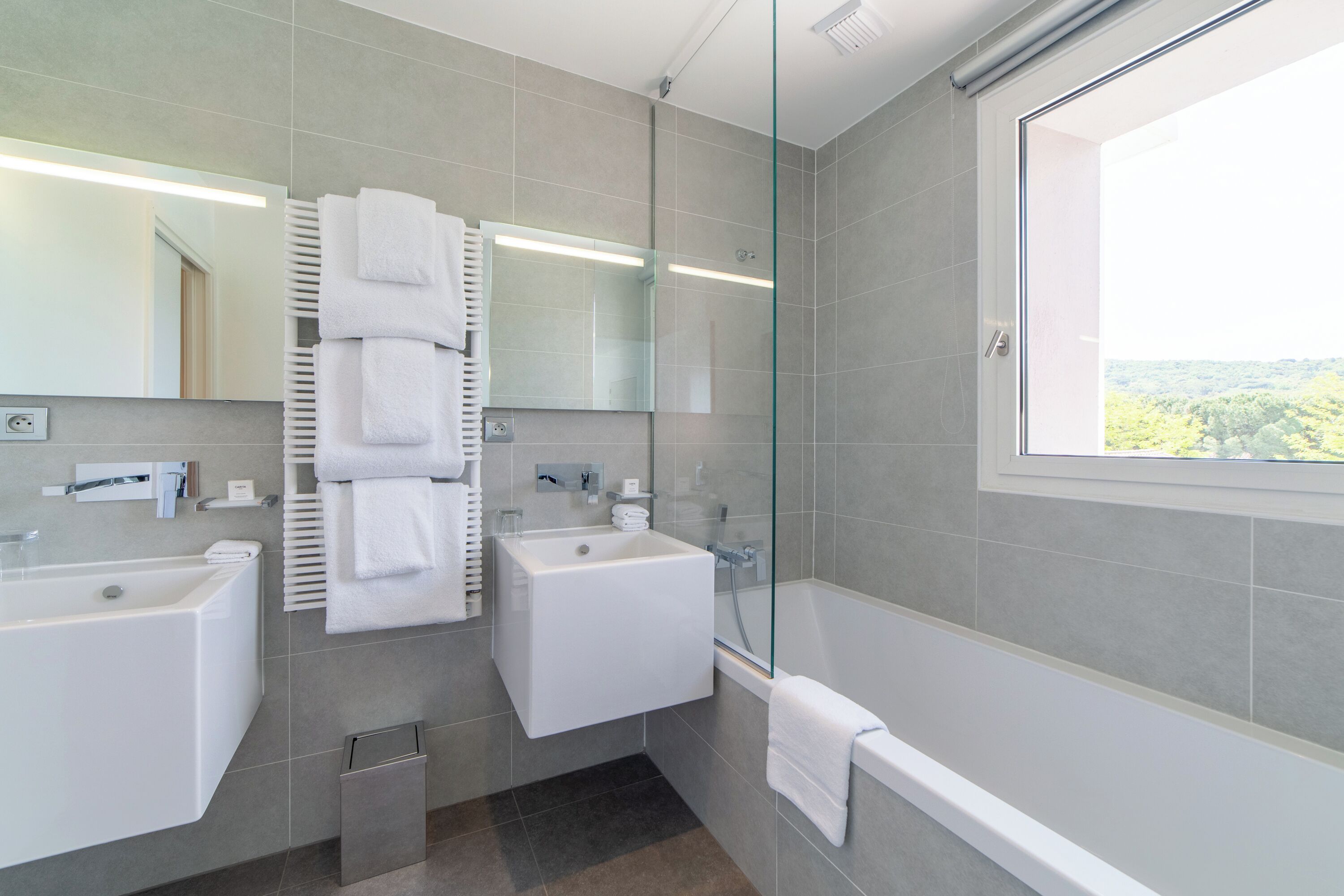 A modern bathroom with two sinks, a glass-enclosed shower, and a large window offering a view.