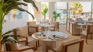 Een ruim, goed verlicht eetgedeelte met ronde tafels en comfortabele stoelen, omgeven door grote ramen met uitzicht op palmbomen en een gebouw.