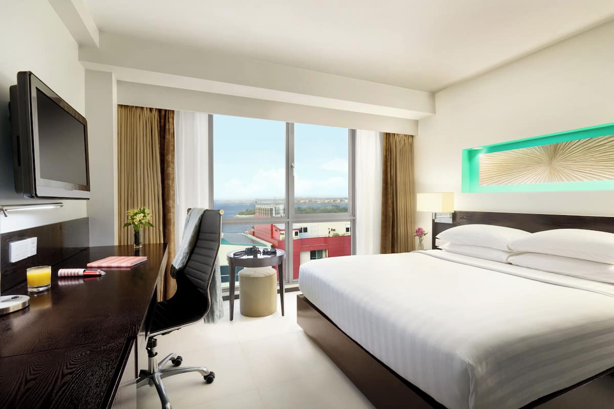 A hotel room with a bed, a desk with a TV, a chair, a small table, and a view of the city through large windows.