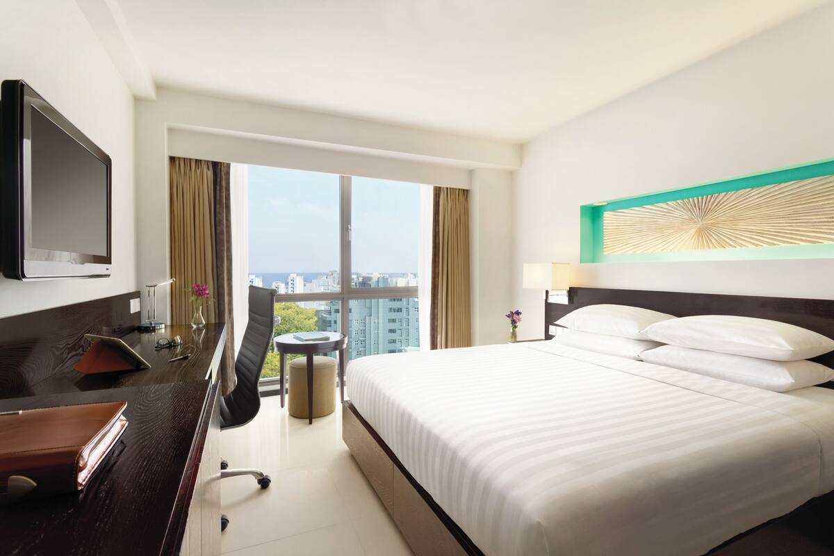 A hotel room with a large bed, a desk with a TV, a chair, and a view of the city through large windows.
