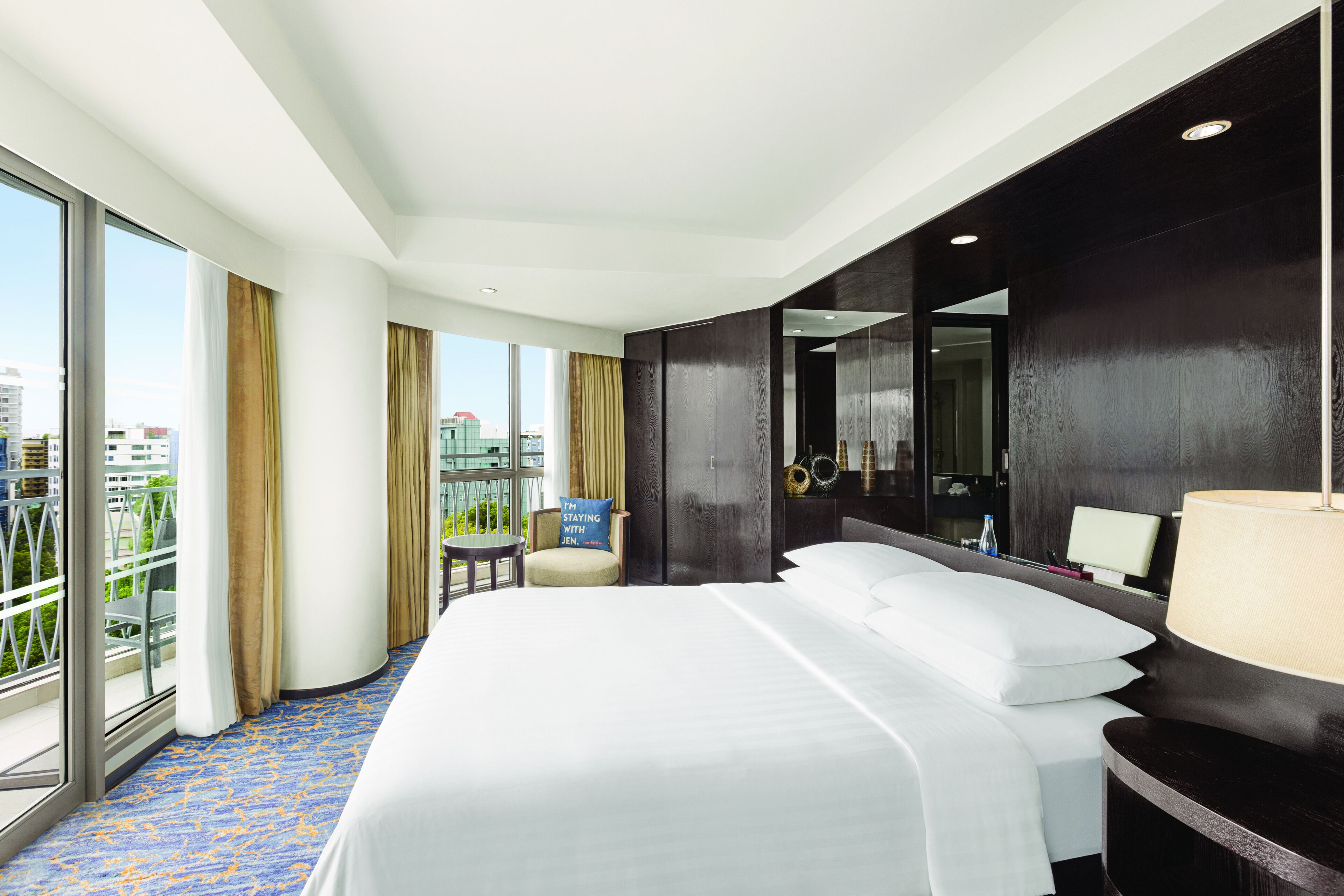 Habitación de hotel con una cama grande, un escritorio, una silla, un balcón con vistas a la ciudad y un ventanal amplio.