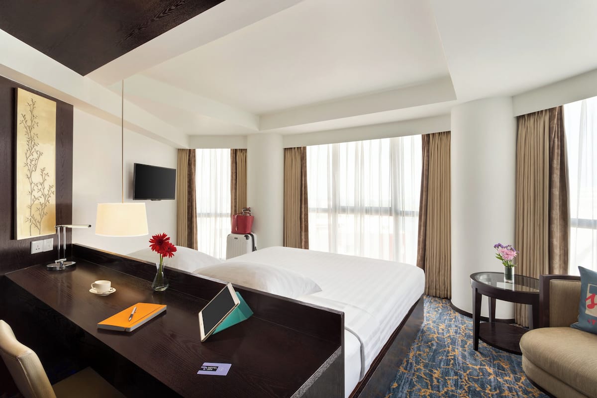 A hotel room with a bed, desk, chair, TV, and large windows with curtains.