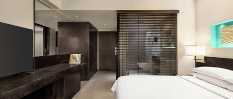 Ein modernes Hotelzimmer mit einem großen Bett, einem Schreibtisch, einem Spiegel und einem durch eine Schiebetür einsehbarer Badezimmer.