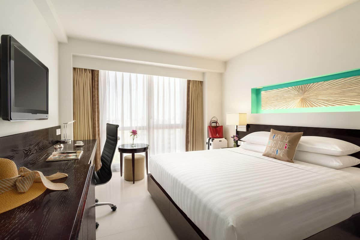 A hotel room with a bed, a desk with a TV, a chair, a small table with a flower, and a window with curtains.