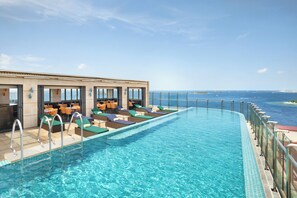 Un espace piscine doté de chaises longues et de tables, une vue dégagée sur l’océan et un bâtiment avec de grandes fenêtres.