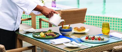 A dining table with a meal, a glass of juice, and a view of a pool.